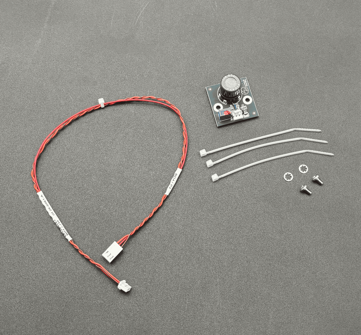 A small electronic control module kit on a dark gray textured background, including a red and white wire harness with a connector, a small circuit board with a rotary knob, three white zip ties, two screws, and two washers.